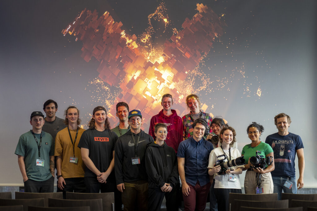 A group picture with Axel Bauer in the first row wearing a blue shirt and red pants and his director headset. Behind a projection with the Expanded Animation 2022 Logo is visible.