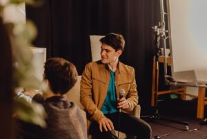 Axel Bauer sitting at the set and looking to the left at the (non visible) interviewer during the livestream. He holds a microfone in his left hand and wears a brown jacket. Behind, set decorations such as vases inside a shelf, lights and a black curtain is visible.