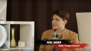 Axel Bauer sitting at the set and looking to the left at the (non visible) interviewer during the livestream. He holds a microfone in his left hand and wears a brown jacket. Behind, set decorations such as vases inside a shelf, lights and a black curtain is visible.