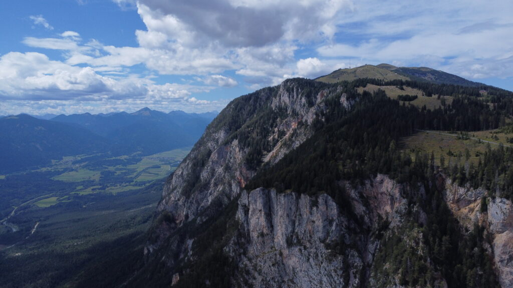 A big mountain next to a valley in summer times. Sharp slopes gently merge into wooded ridges.