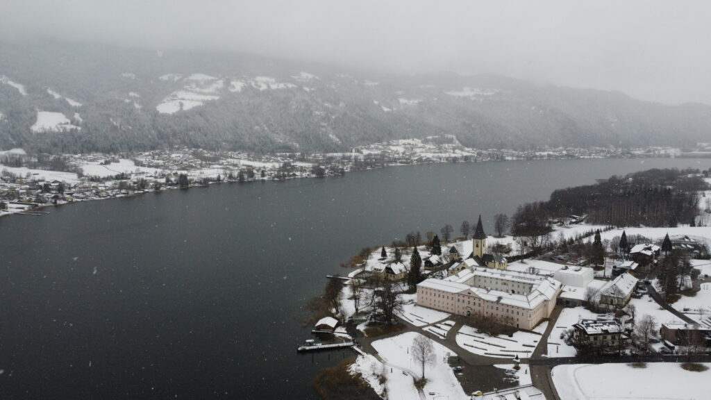 A snow covered diocese with a church next to a lake shot from a drone while flying.