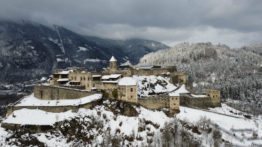 A picture of a castle which was shot on a drone while flying during winter times. Snow covers the roofs and trees. Behind the castle, there is a snow covered mountain.