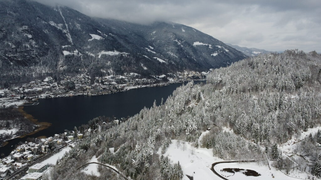 A picture of a lake behind a snow covered mountain, which was shot on a drone while flying during winter times. Behind the lake is a even bigger moutain located.